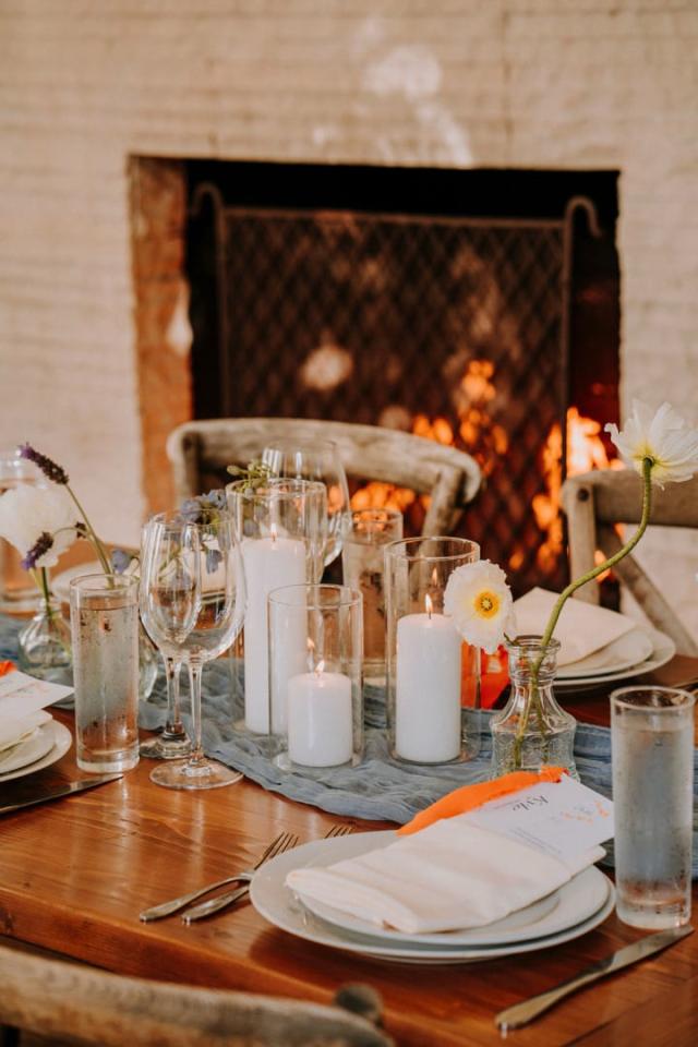 Mid shot of wooden table, place sets, candles, flowers, and fire place in the background for Veronica & Jake’s Wedding