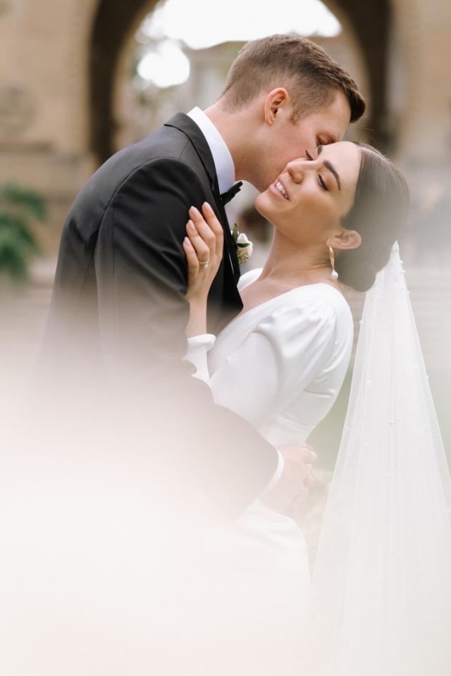 A bride and groom sharing a tender kiss. the bride is in a white dress with a long veil, and the groom is in a black suit. the image captures a romantic moment, with soft focus in a historical setting.