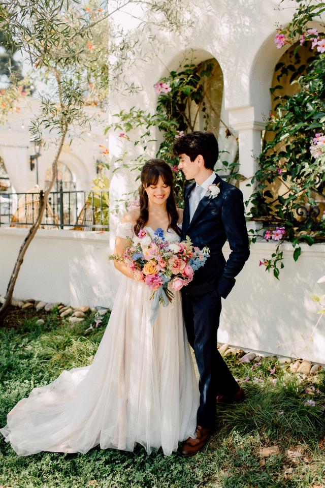 A bride and groom stand together outdoors, surrounded by flowering plants. The bride holds a colorful bouquet, and the groom is dressed in a dark suit.