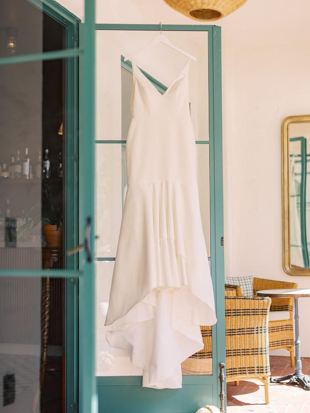 A white sleeveless dress with a fitted bodice and flared hem hangs on a hanger from a glass door in a sunlit room.