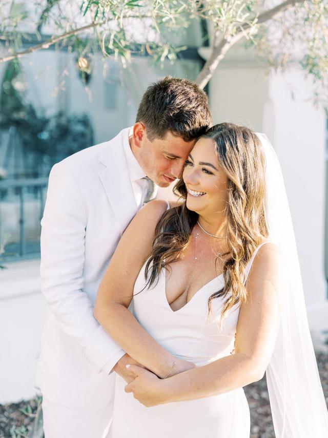 A bride and groom in white attire embrace and smile outdoors, with greenery and a white building in the background.