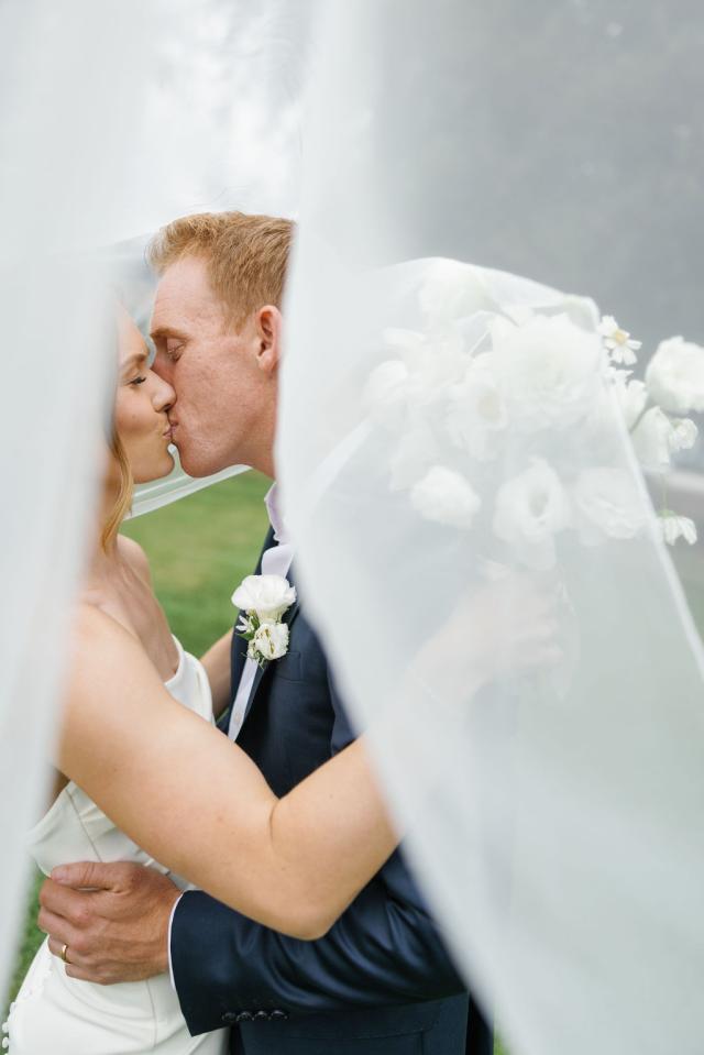 A bride and groom kiss outdoors beneath a sheer white veil, with the bride holding a bouquet of white flowers.