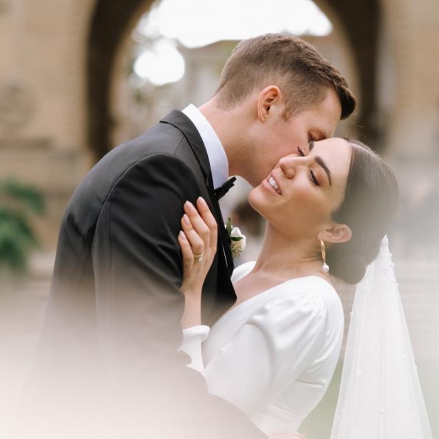 A bride and groom sharing a tender kiss. the bride is in a white dress with a long veil, and the groom is in a black suit. the image captures a romantic moment, with soft focus in a historical setting.