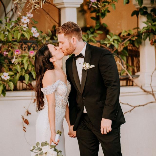 Bride in an off-shoulder lace gown and groom in a dark suit kiss, holding a bouquet, with a floral adorned wall behind.