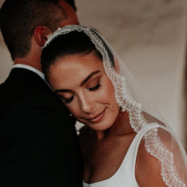 Bride with a lace veil gently rests her head on the groom's shoulder in a tender, intimate moment.