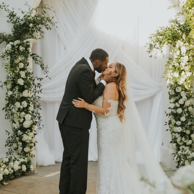 A bride and groom kissing at an outdoor altar adorned with white drapes and white floral arrangements.