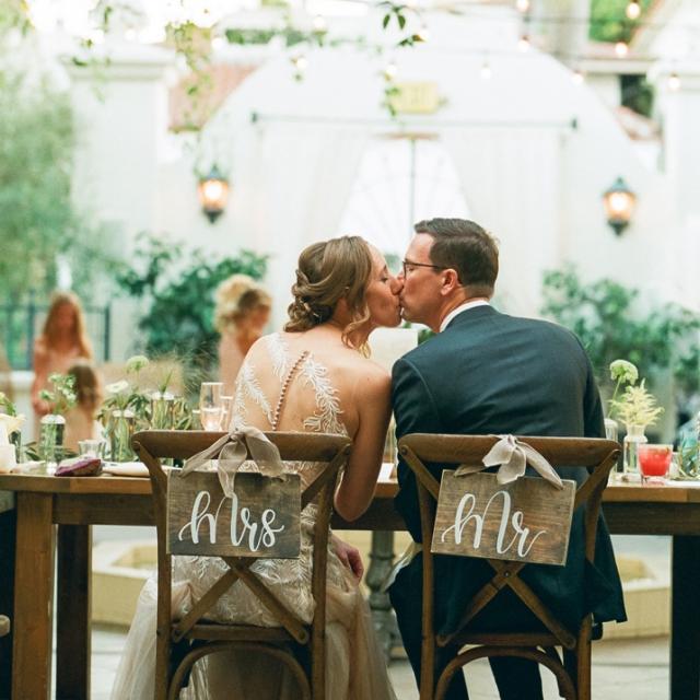 A bride and groom kiss at a decorated table labeled with "mrs." and "mr." signs, surrounded by a warmly lit garden setting.