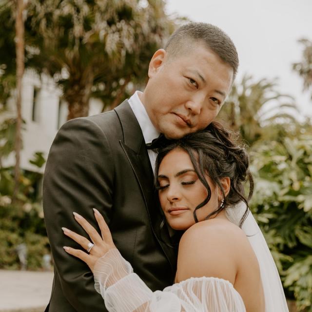 A bride and groom embrace lovingly, the groom in a black suit and the bride in a white dress with billowing sleeves, with soft focus greenery in the background.