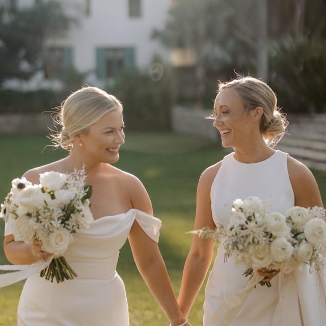 Two women in elegant white dresses holding hands and bouquets, smiling at each other in a garden setting.