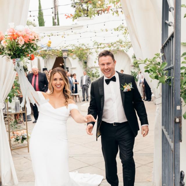 A radiant bride in a white gown and a groom in a black tuxedo holding hands joyfully exit through a gate, with wedding guests in the background.