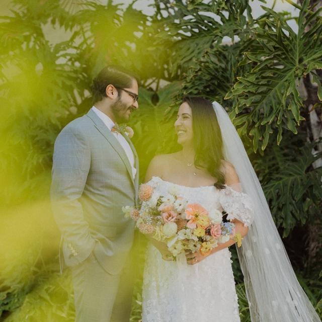A bride and groom smiling at each other in a lush garden, with the bride holding a bouquet and the groom in a light gray suit.