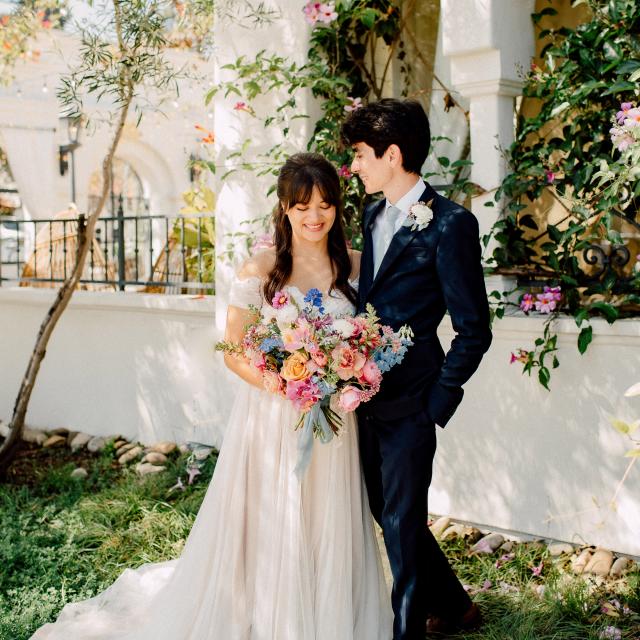 A bride and groom stand together outdoors, surrounded by flowering plants. The bride holds a colorful bouquet, and the groom is dressed in a dark suit.