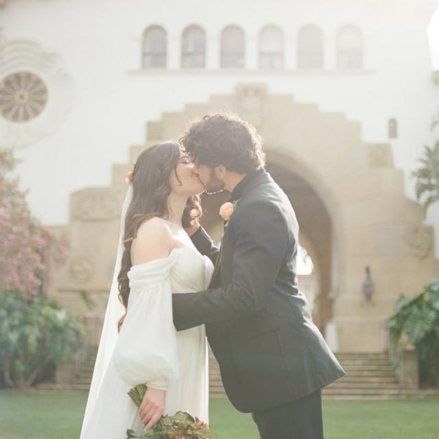 A bride and groom share a kiss outside a grand building. The bride holds a bouquet of orange flowers, and both are dressed in formal attire.