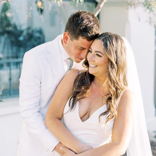A bride and groom in white attire embrace and smile outdoors, with greenery and a white building in the background.