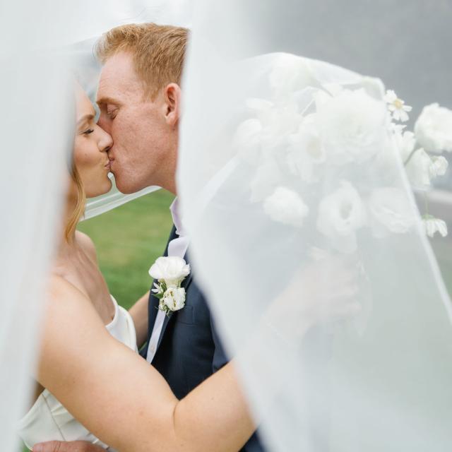 A bride and groom kiss outdoors beneath a sheer white veil, with the bride holding a bouquet of white flowers.