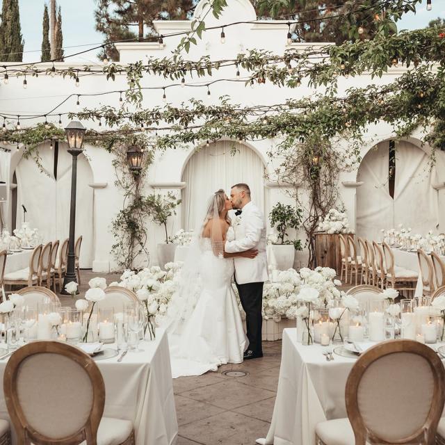 A bride and groom kiss in a candlelit outdoor wedding reception with white floral decor.
