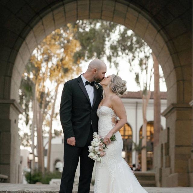 A bride and groom share a kiss under a stone archway on steps, with the bride holding a bouquet.