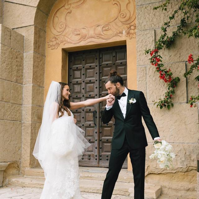 A groom in a tuxedo kisses the hand of a smiling bride in a white dress outside an ornate stone building.