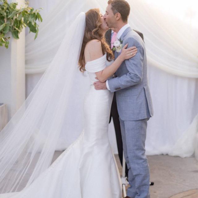 Bride and groom share a kiss during an outdoor wedding ceremony with draped white fabric backdrop.